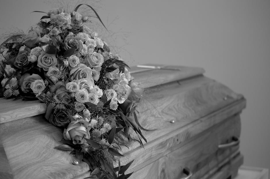A large, elegant funeral floral arrangement resting on the closed lid of a wooden casket. The bouquet comprises numerous fresh roses in shades of white and pink, interspersed with smaller filler flowe
