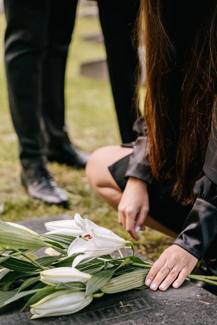 A person dressed in black kneels on the ground, carefully arranging a floral bouquet consisting of large white lilies with open petals and prominent stamens, surrounded by long, green leaves. The bouq