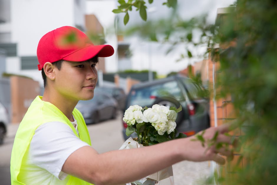 A young male florist dressed in a white shirt, high-visibility yellow vest, and red cap is arranging a bouquet of fresh white flowers, possibly hydrangeas, near a brick wall on a street with parked ca