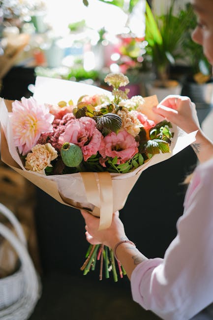 A person holding a large bouquet of fresh flowers, including pink peonies, white and purple blooms, and lush green foliage, wrapped in light beige paper with cream-colored ribbon, likely for a floral 