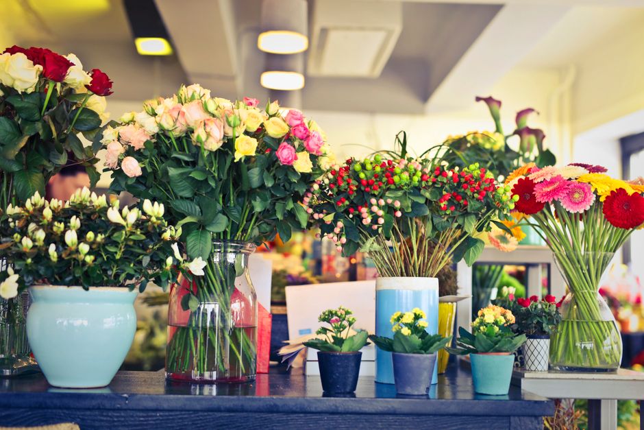 A variety of fresh floral arrangements and bouquets displayed on a wooden table in a flower shop, including a large bouquet of pink and yellow roses in a clear glass vase, a smaller arrangement of whi