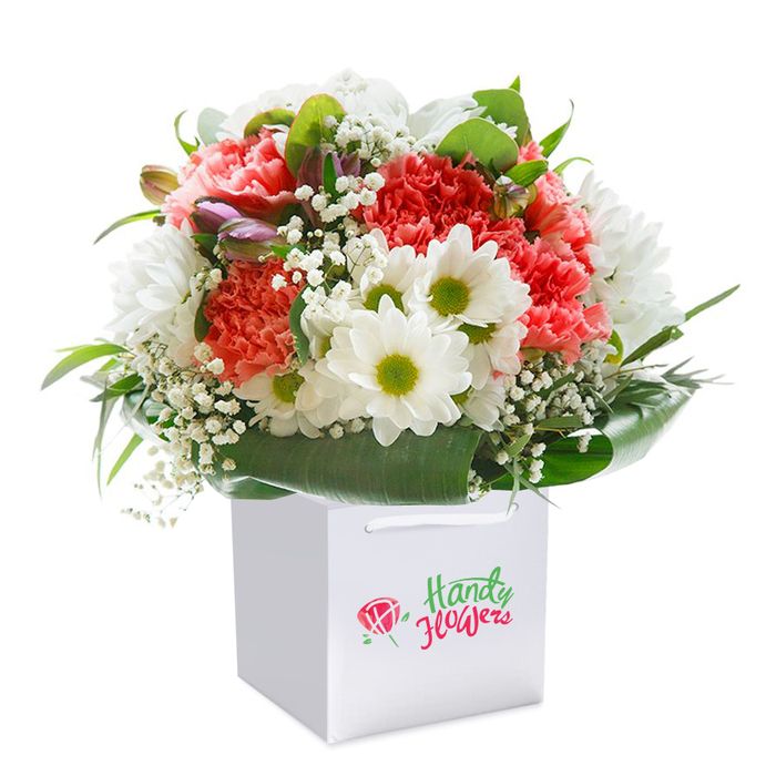 Set on a flawless white background, a vibrant floral arrangement stands upright in a square white gift bag, offering the perfect embodiment of Keston's natural charm and local floral artistry. The bouquet dazzles with soft coral-pink and rich red carnations at its heart, each flower's ruffled, frilled petals contributing both richness and a welcoming softness, reminiscent of Keston's blooming spring gardens. Bright white daisy-like blooms, with striking chartreuse-tinged centers, introduce cheerful contrast and freshness. Weaving through this palette, delicate sprigs of white baby's breath provide airy lightness, reflecting the gentle breezes of Keston Common on a mild morning. Lush, glossy green foliage with broad leaves cradles the base-creating a natural collar that frames the arrangement with elegance and structure, echoing the well-kept hedgerows seen along Heathfield Road. A scattering of closed, purple-hued buds-perhaps a nod to the first hint of wild violets in local parklands-contribute intrigue and visual depth. The robust white bag, topped with satin ribbon handles, hints at careful delivery and thoughtfulness, whether for a birthday, anniversary, or spontaneous gesture. The detailed composition, fullness, and gleaming freshness are all a testament to the knowledge and artistry found in Keston's beloved flower shops-each stem arranged to celebrate life's meaningful moments in this close-knit community.